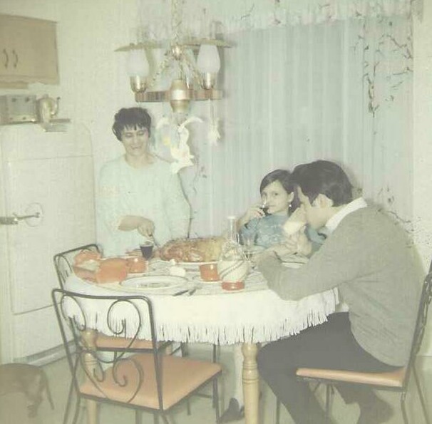 A woman stands behind a round dining table in a vintage kitchen, serving while a man and a girl sit and share a meal with a roasted chicken and dishes on the table.