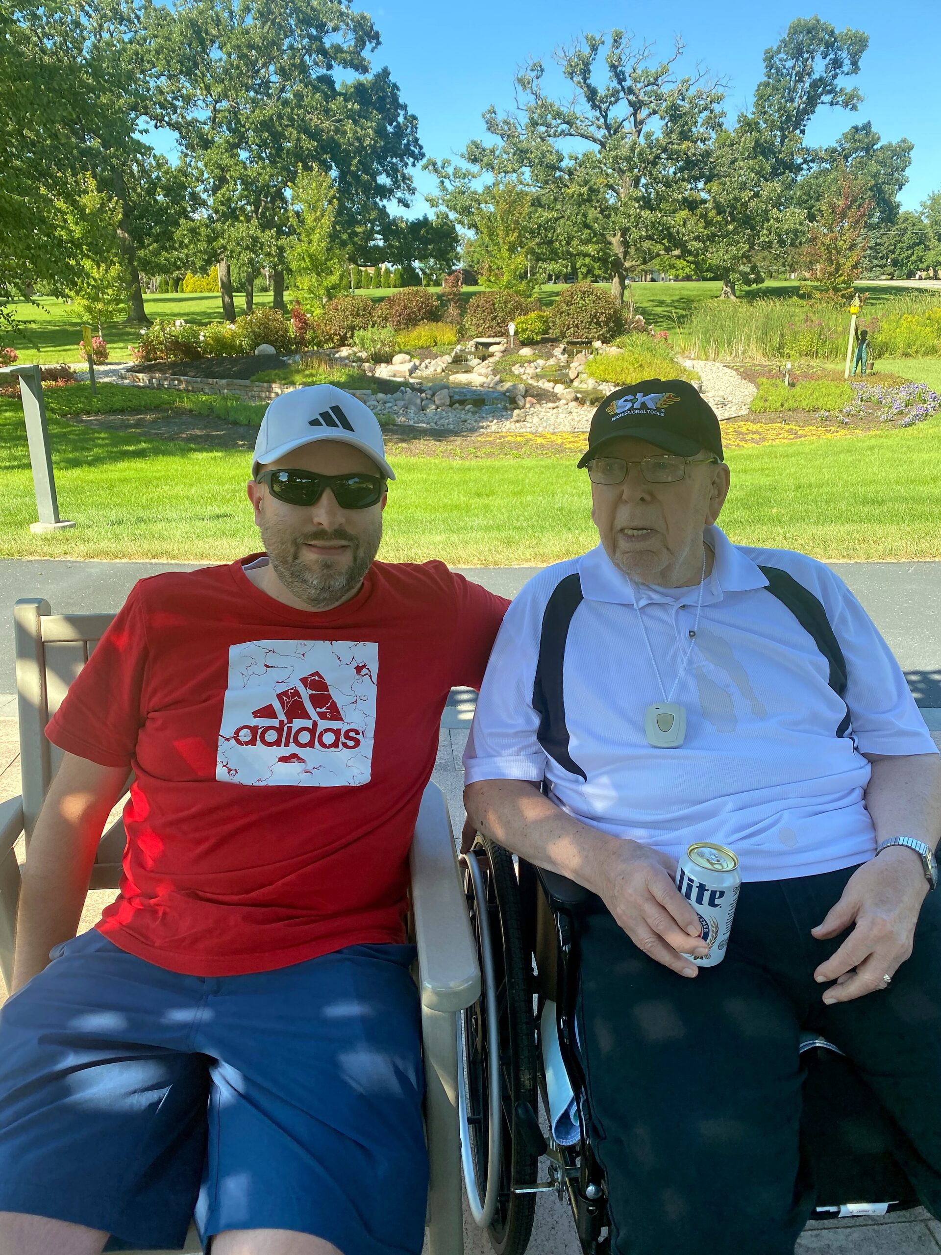 Two men sit on a park bench on a sunny day; one wears a red Adidas shirt and white cap, the other is in a wheelchair with a white polo and black hat, holding a beer can.