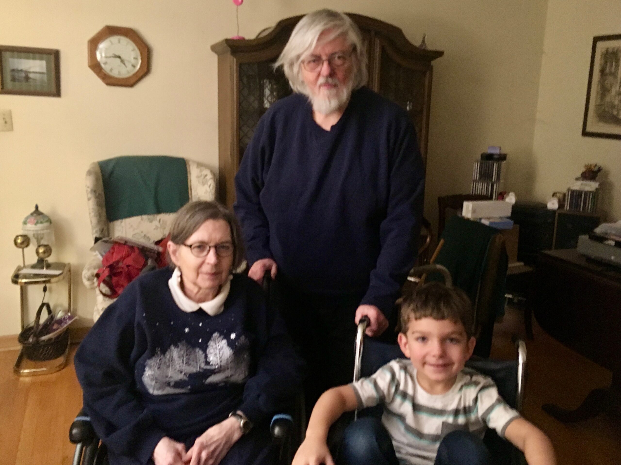 Three-generation family in a living room: an elderly man with white hair stands behind a seated woman and a smiling boy in a wheelchair in the foreground.
