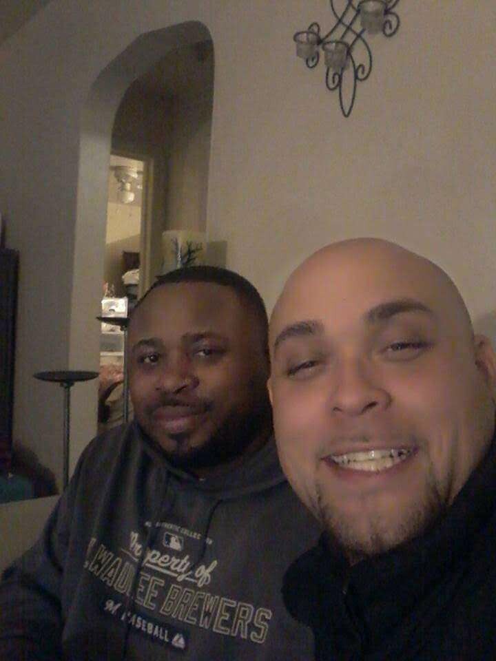 Two men posing for a selfie indoors, smiling at the camera in a living room setting.