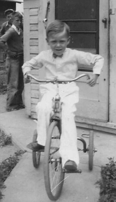 Young boy in light clothes rides a tricycle on a sidewalk in front of a porch, while two other kids stand nearby.