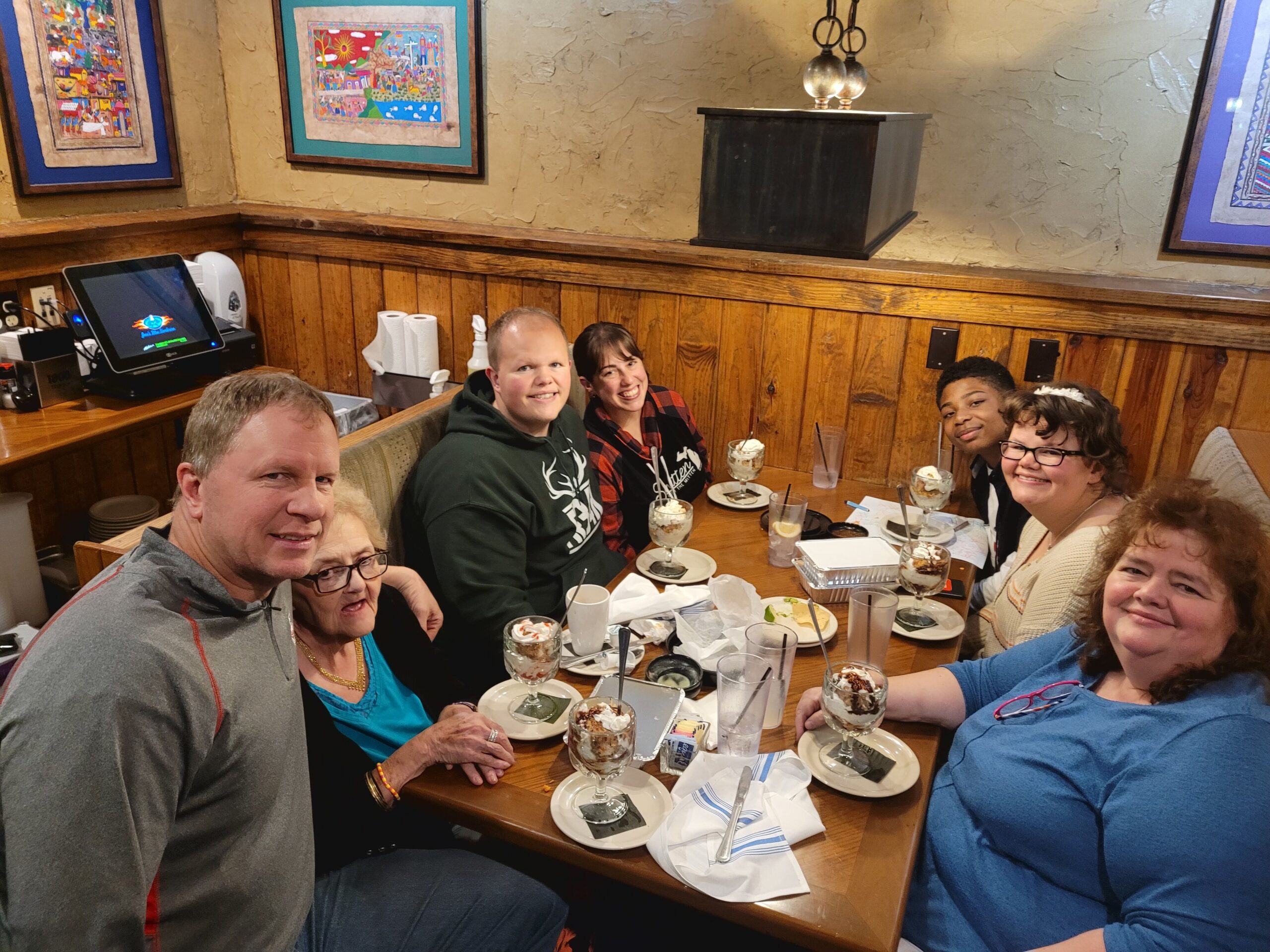 Group of friends and family gathered at a restaurant table, posing for a selfie with desserts and milkshakes in front of them.