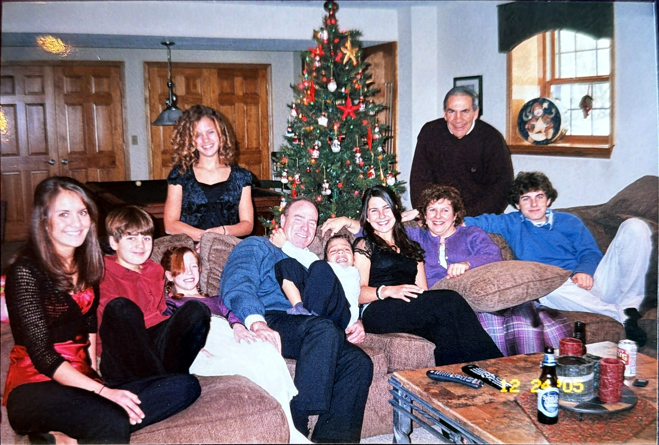 A multi‑generational family sits on a living room couch around a decorated Christmas tree, smiling at the camera.