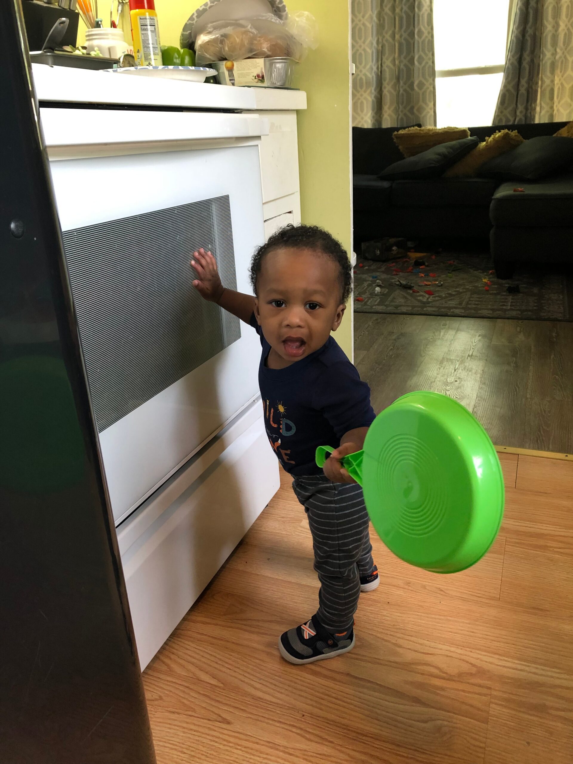 Toddler stands by a white oven in a kitchen, holding a bright green plastic plate and looking at the camera.