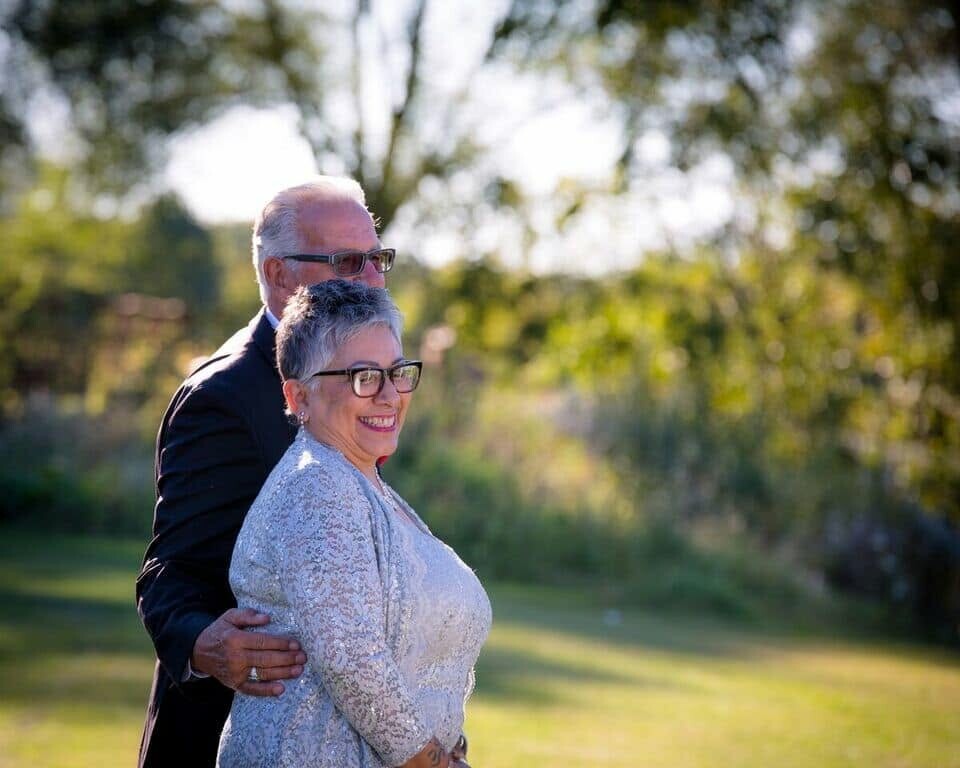 Smiling couple outdoors, man embracing woman from behind in a sunlit park.
