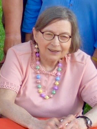 Elderly woman wearing a pink top and colorful bead necklace, smiling outdoors.