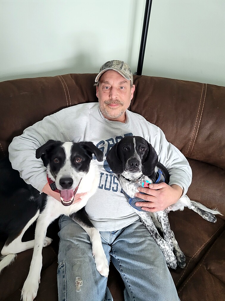 Man in a gray sweatshirt and camo cap sits on a brown couch with two dogs snuggled beside him.