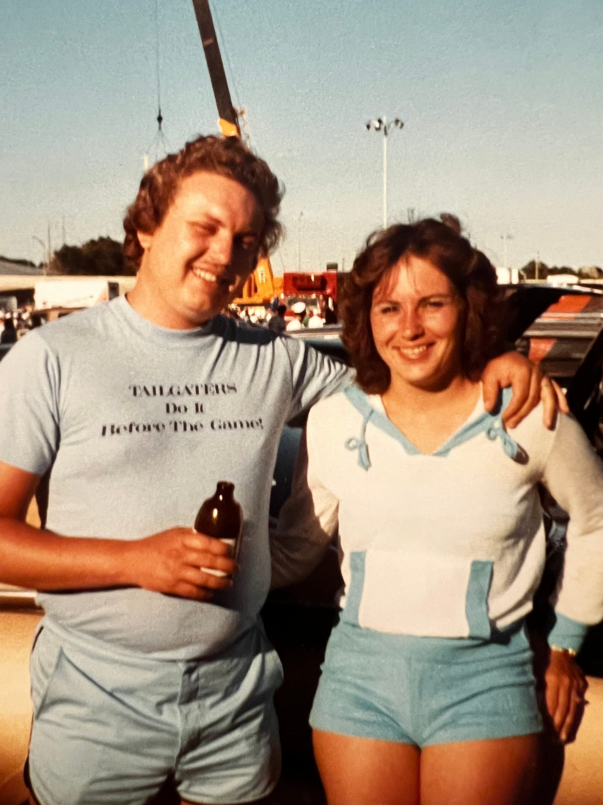 Two adults smiling outdoors, the man holding a beer bottle, with a crane and festival crowd in the background