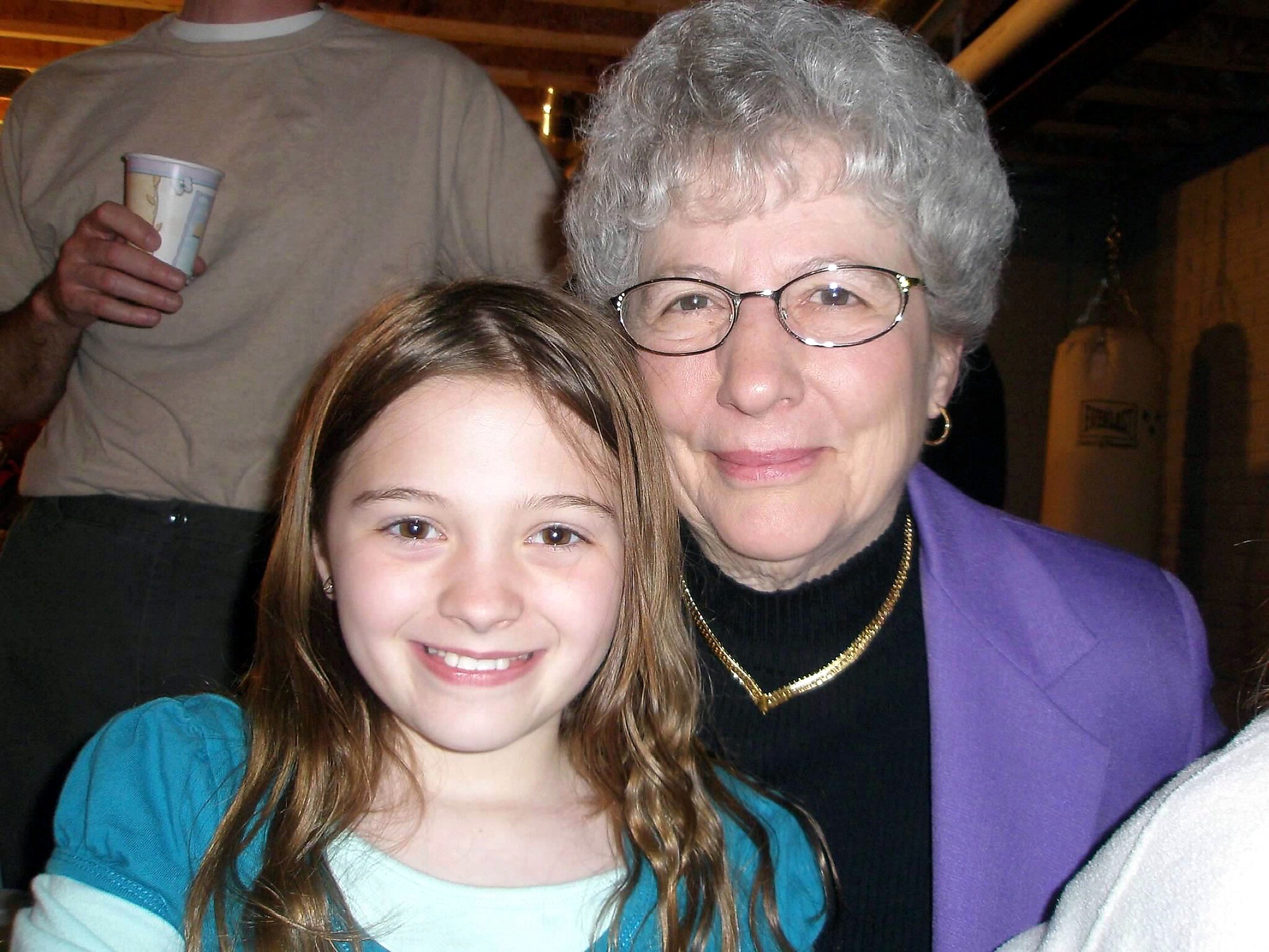 Smiling young girl in a teal shirt posing with an elderly woman wearing glasses and a purple jacket in a casual setting.