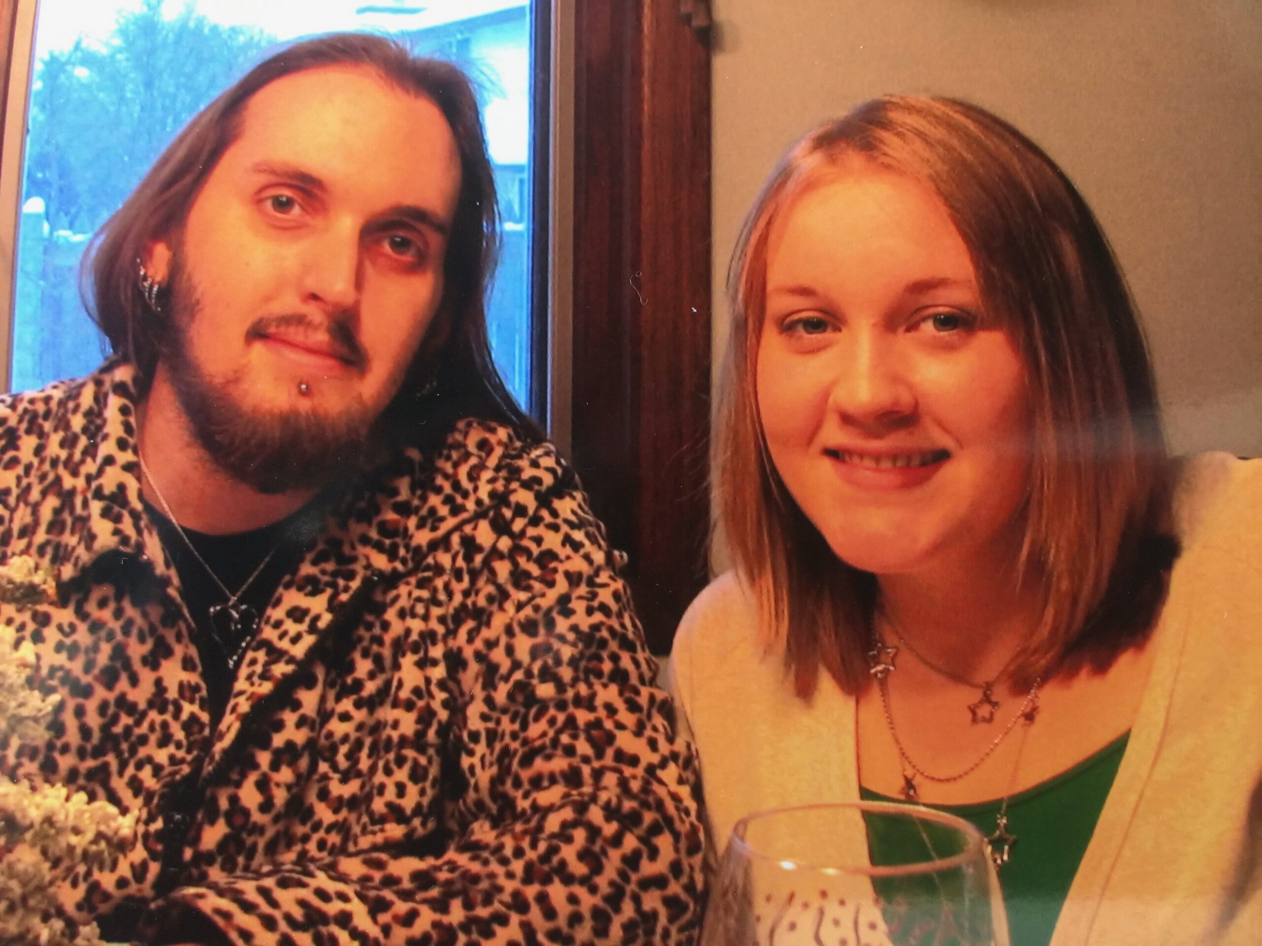 Two friends posing for a party photo: man with leopard-print jacket and lip piercing, woman with blonde hair wearing a green top behind a wine glasss.