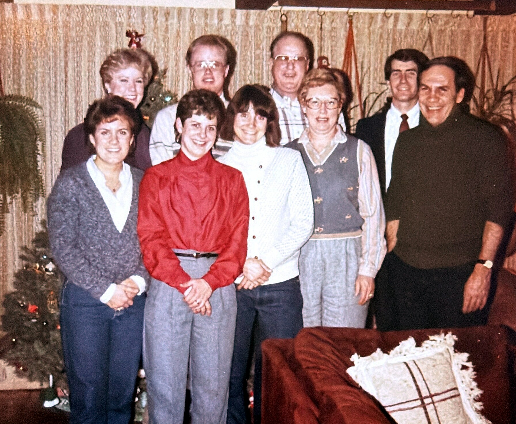 Group of nine adults posing for a photo in a living room, Christmas tree visible on the left and festive decor in the background.