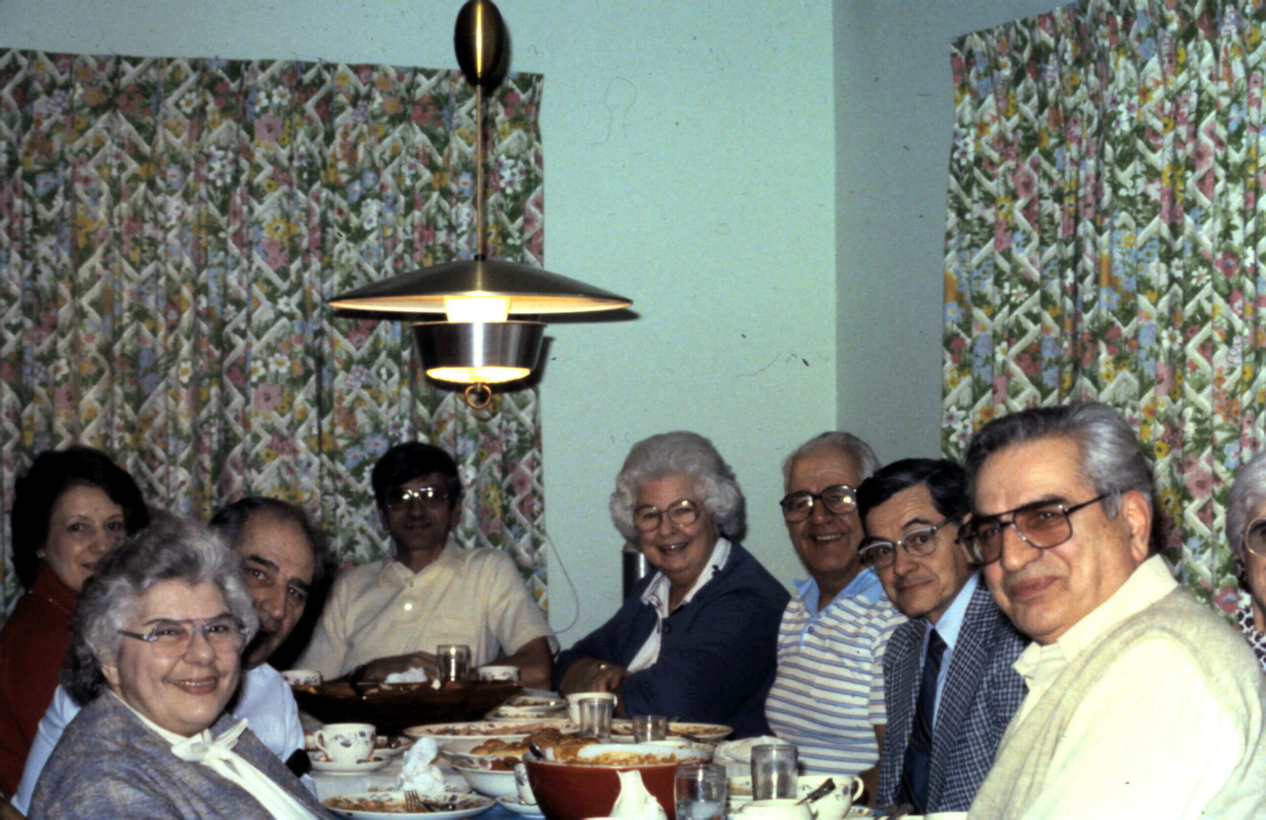 Group of seniors around a dining table, smiling at a meal in a room with floral curtains in the background.