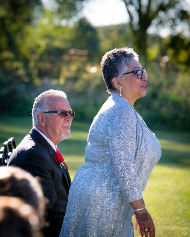 An elderly couple at an outdoor event: a man in a dark suit with a red boutonniere sits beside a woman in a sparkly silver dress, both wearing glasses in a sunny park setting.