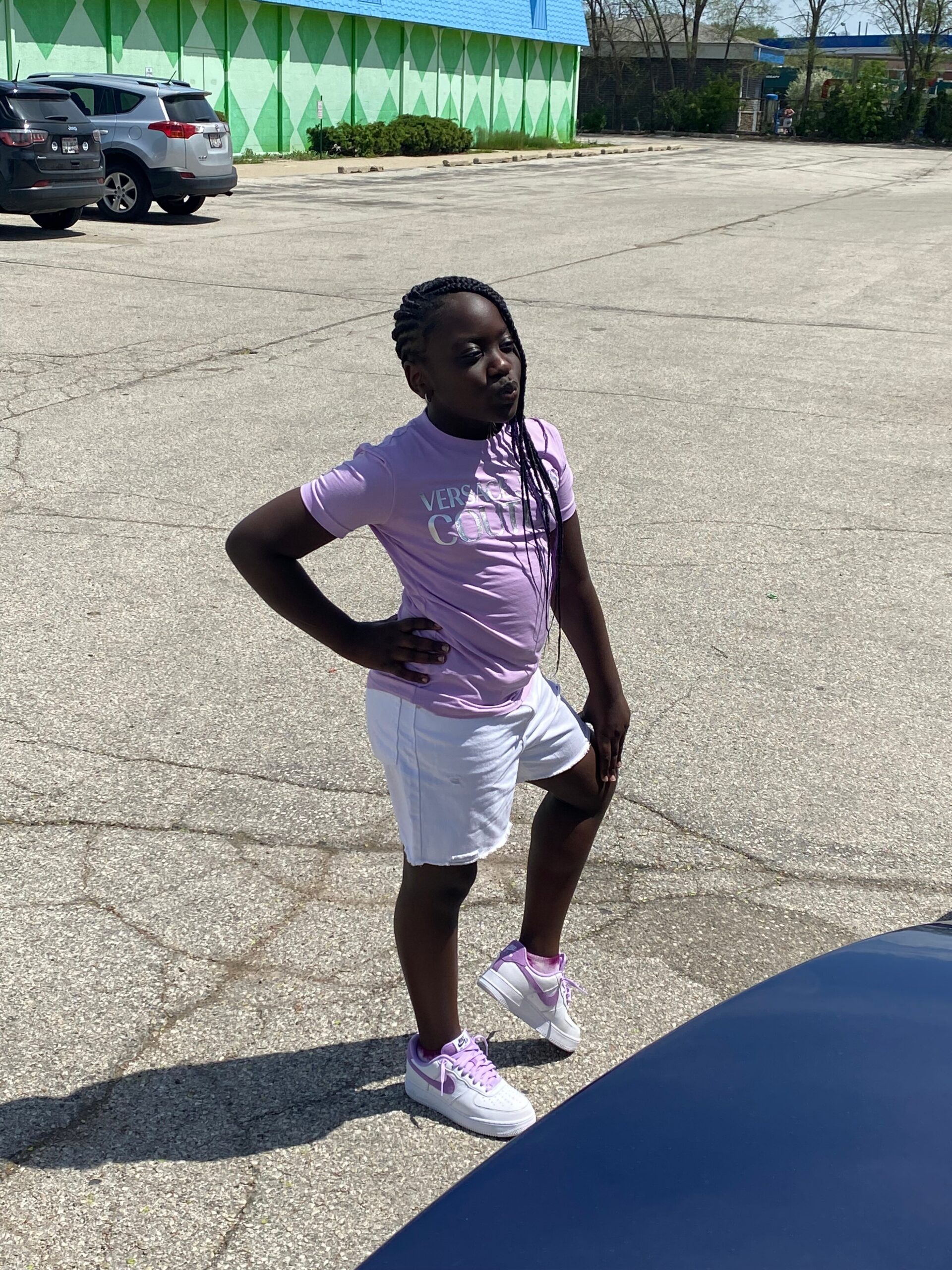 Young girl in a lavender T‑shirt and white shorts poses with hands on her hips in a parking lot, a green diamond-pattern wall behind her.