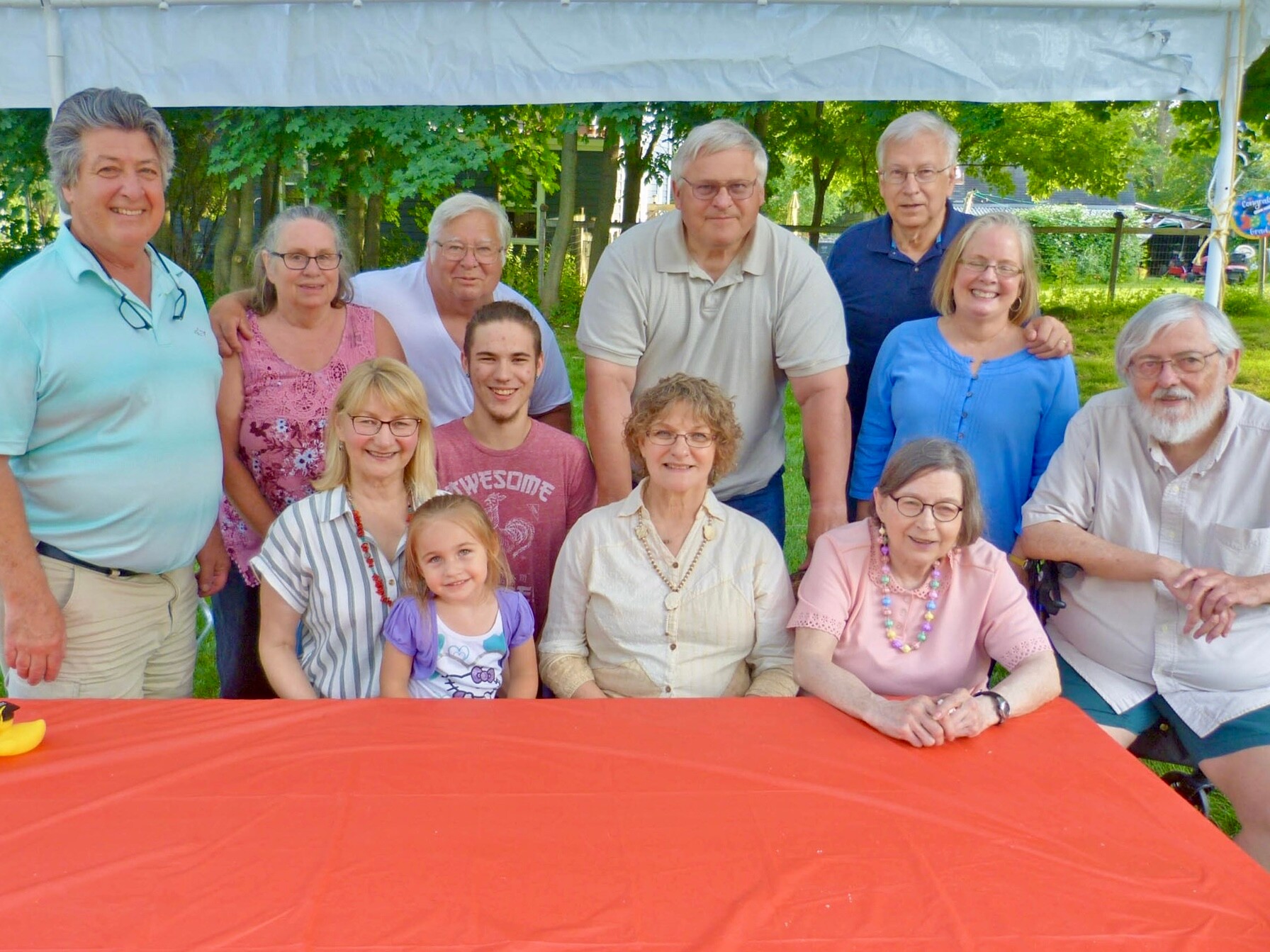 Group of twelve people smiling under a white canopy outdoors, gathered behind a red tablecloth.