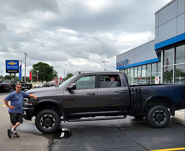 Gray Ram pickup truck parked on a dealership lot, man standing beside the front bumper with another person visible inside the cab