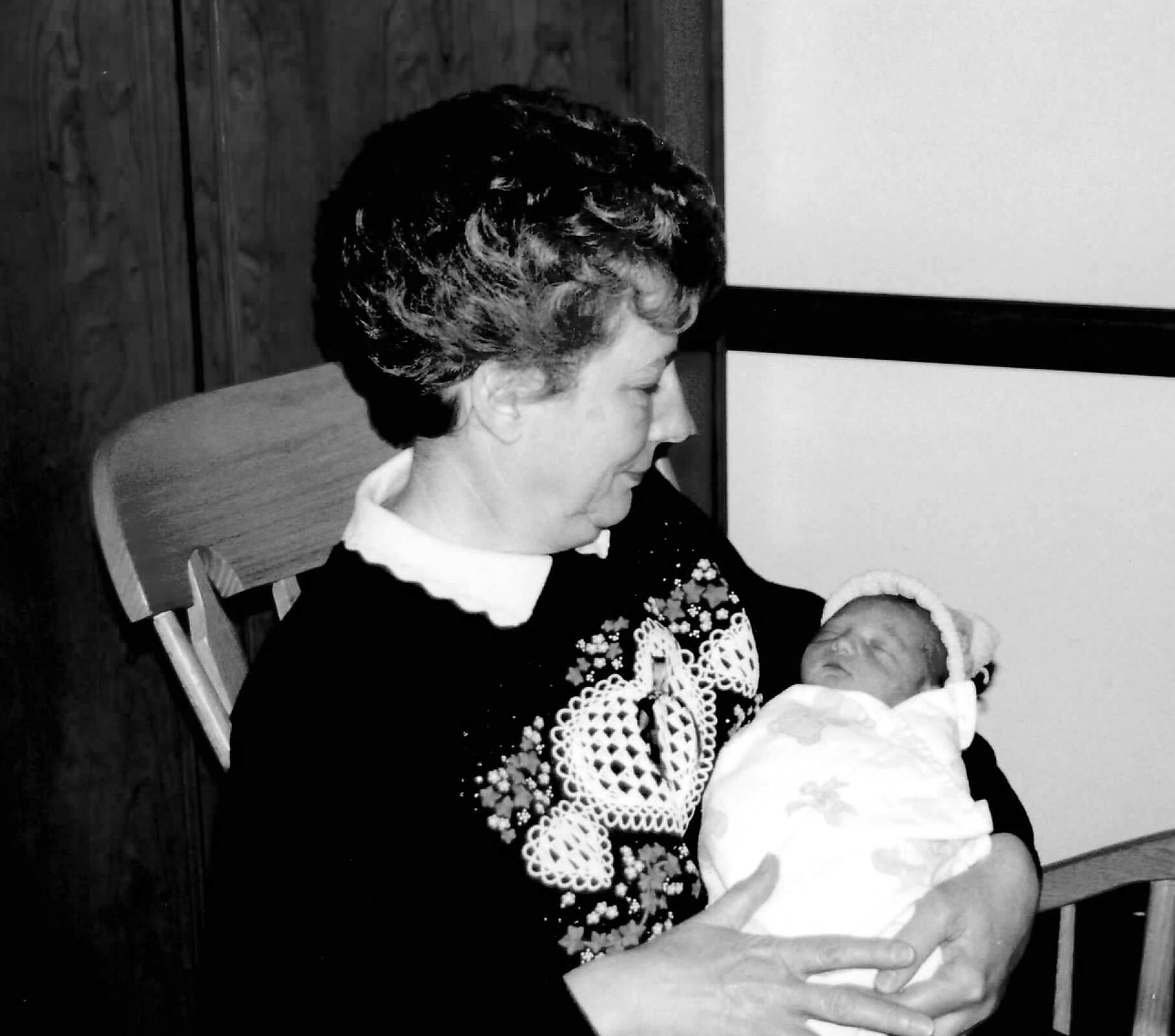 Older woman in a dark dress with lace holding a swaddled newborn baby in her arms, seated in a wooden chair.
