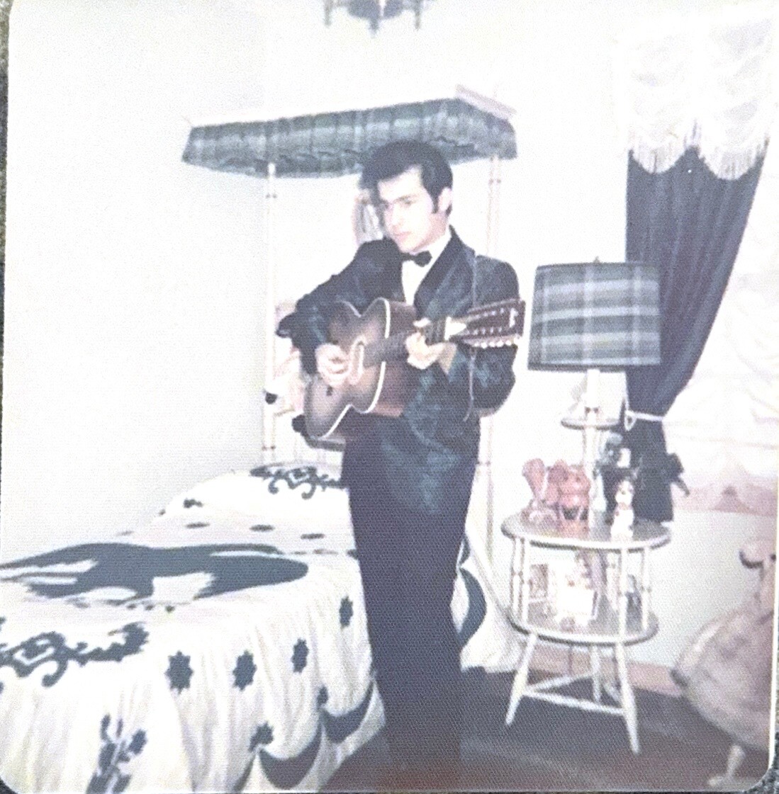 Man in a dark suit playing an acoustic guitar in a vintage bedroom, standing beside a bed and lamp table.