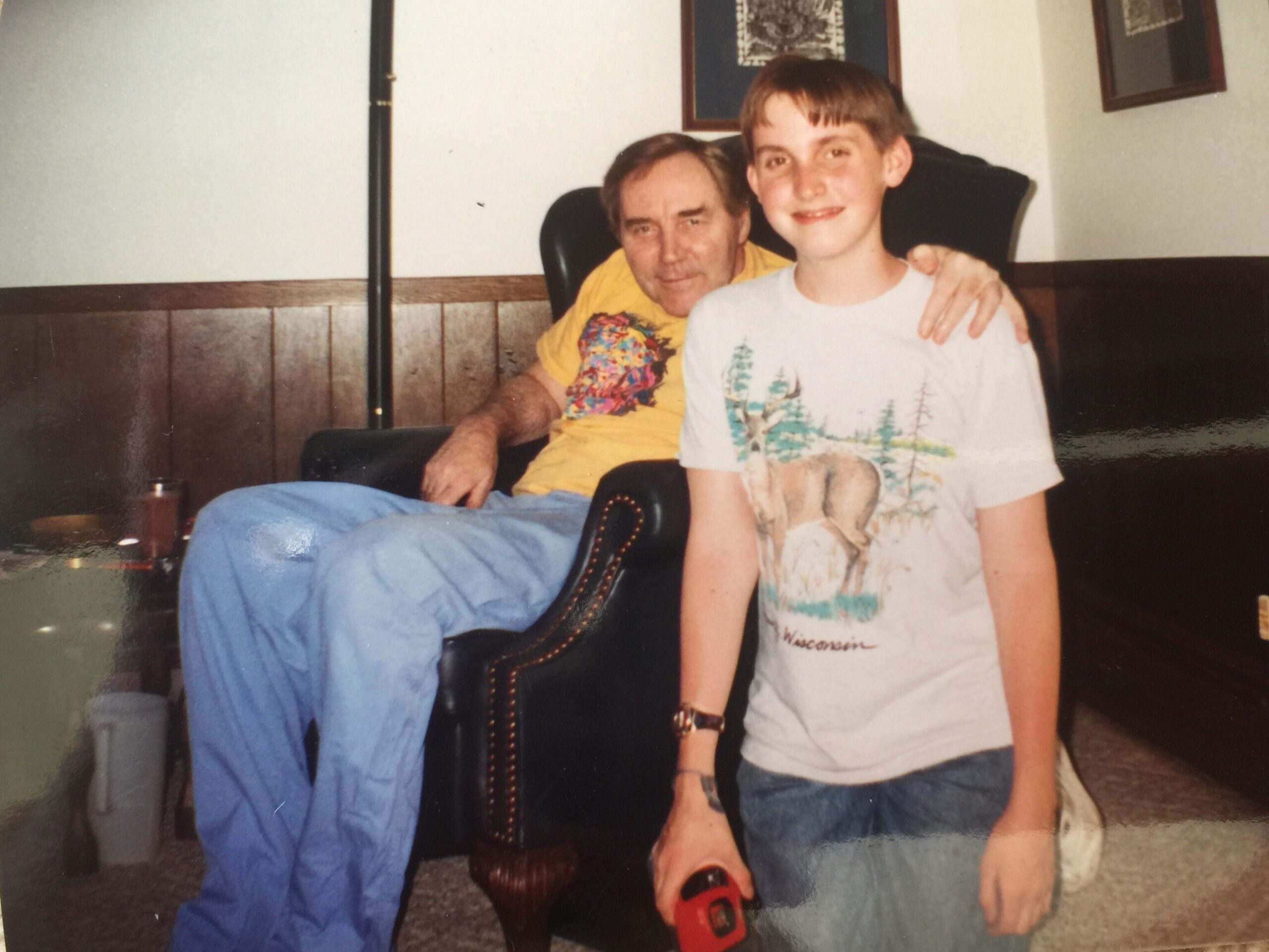 Older man in a yellow shirt sits in a chair with his arm around a smiling boy in a white printed t-shirt, in a wood-paneled living room with framed art behind them.