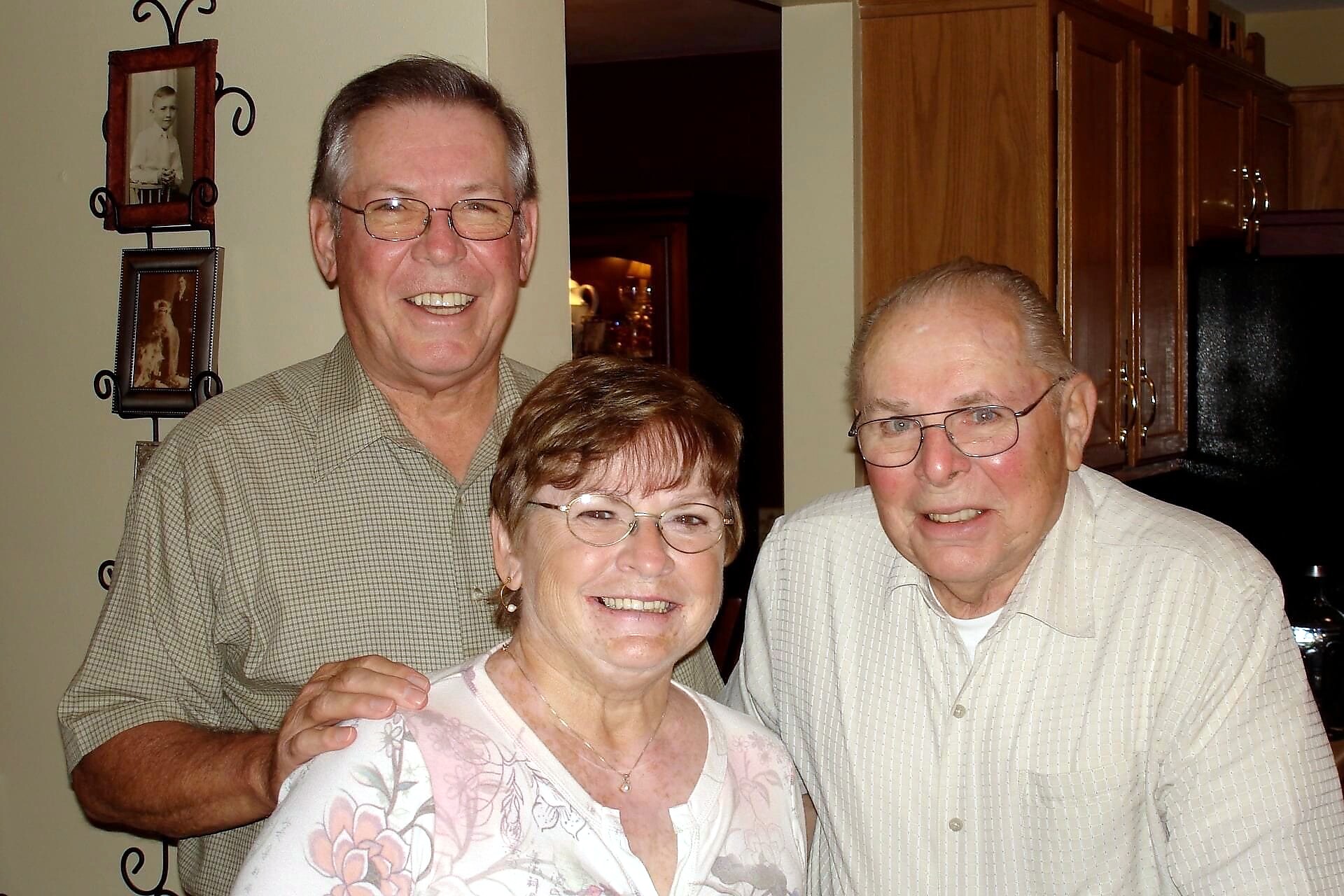 Three smiling seniors pose together in a home kitchen with wooden cabinets in the background.