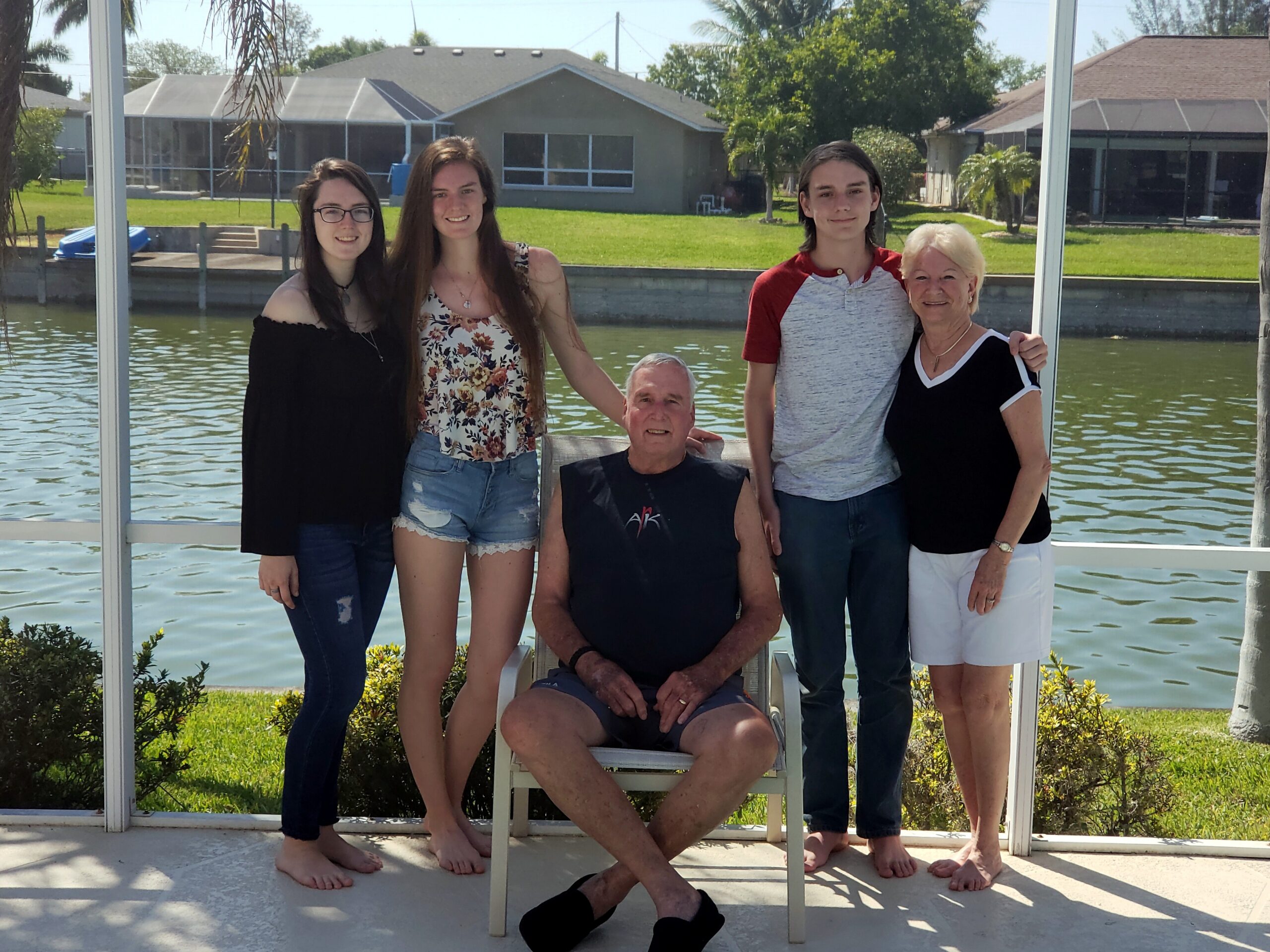 Five people posing outdoors by a canal, with three standing adults and two older adults—one seated in a chair—smiling at the camera on a sunny day.