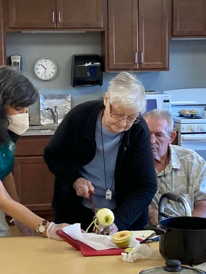 Older woman with short white hair and glasses cuts an apple on a red cutting board while a masked helper assists; a man sits nearby watching in a kitchen with wooden cabinets.