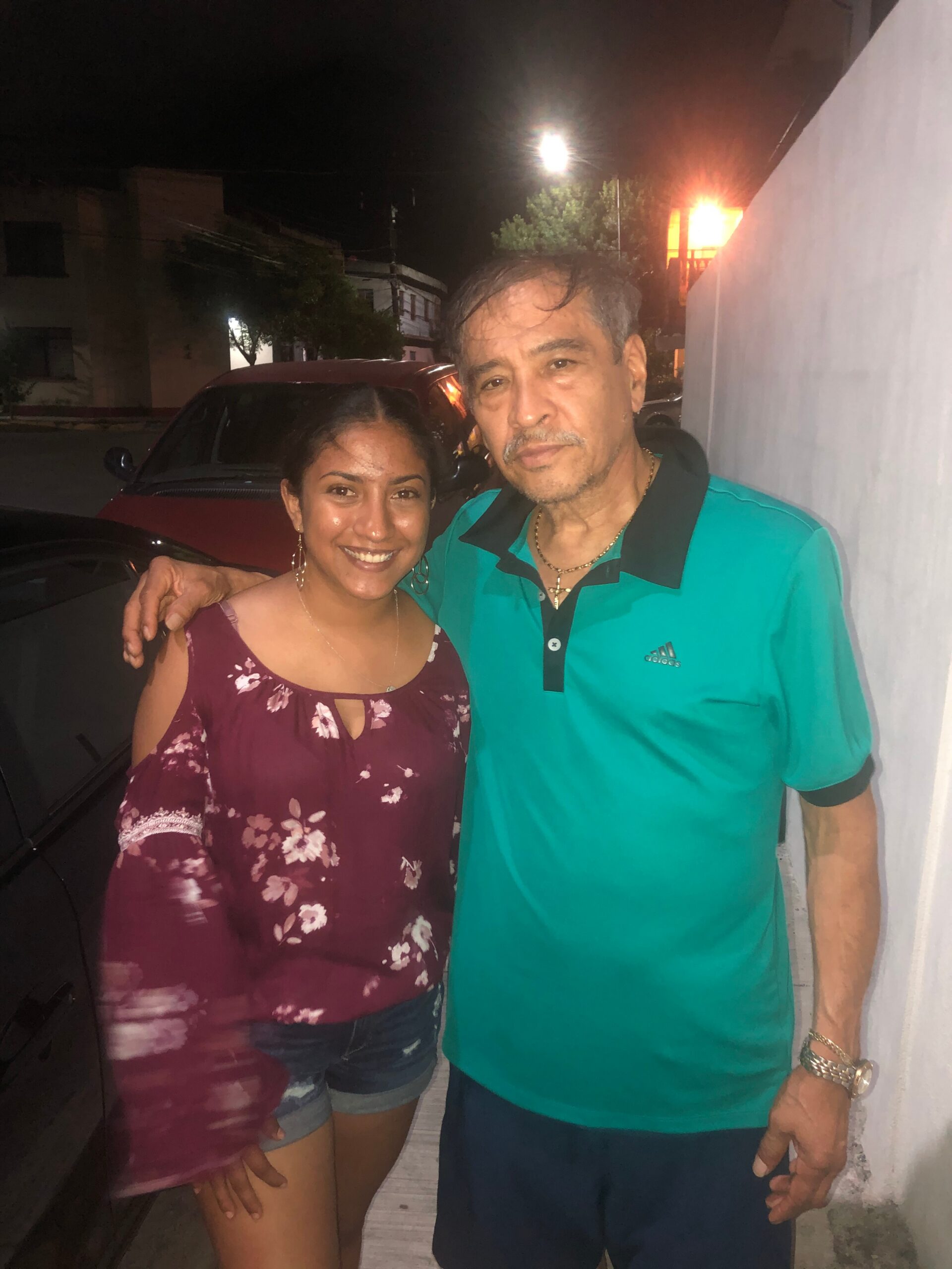 Smiling couple posing together at night outdoors, woman in a maroon floral top and shorts, man in a teal polo shirt next to a white wall and parked car behind them.