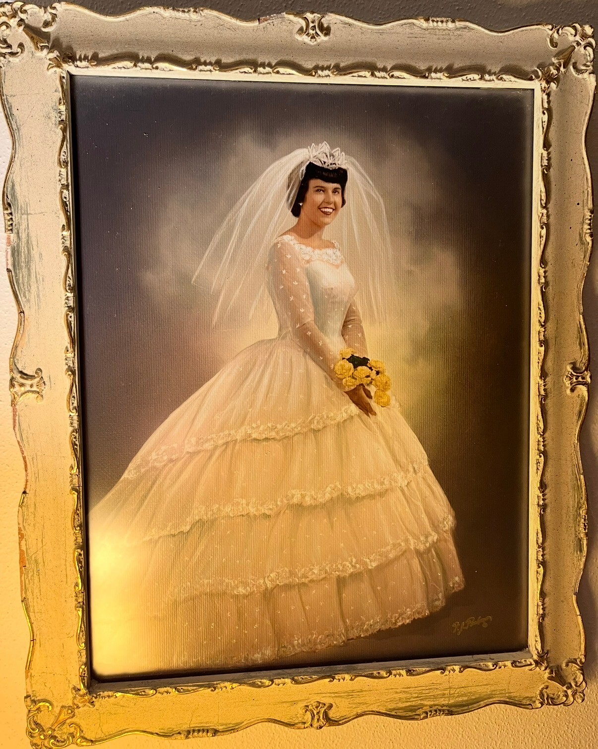 Bride in a vintage white wedding gown with a long veil and crown, holding a small bouquet of yellow roses, posed for a studio portrait in an ornate gold frame