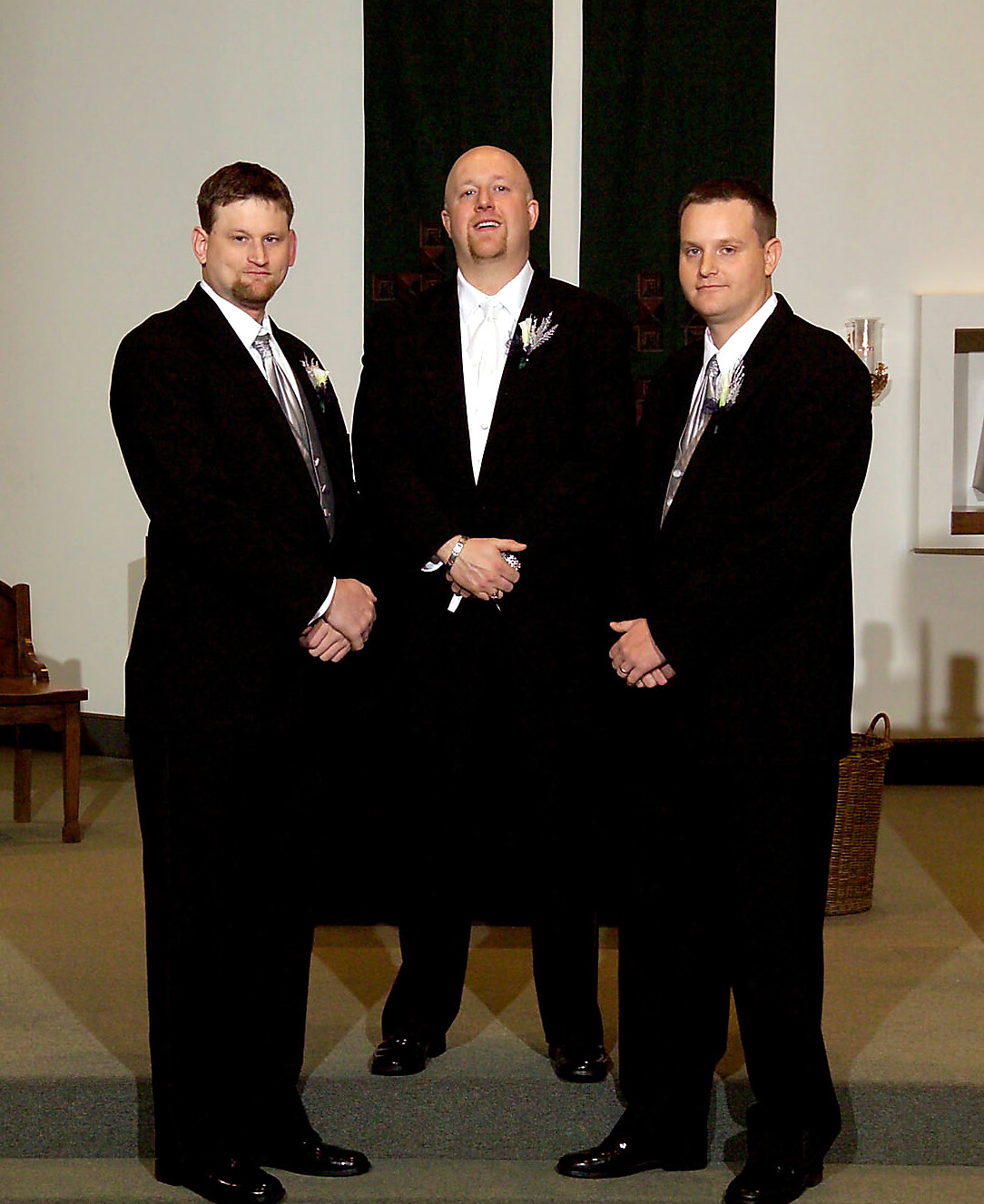 Three men in black suits and white shirts pose together in a church setting, each with a boutonniere on the lapel.