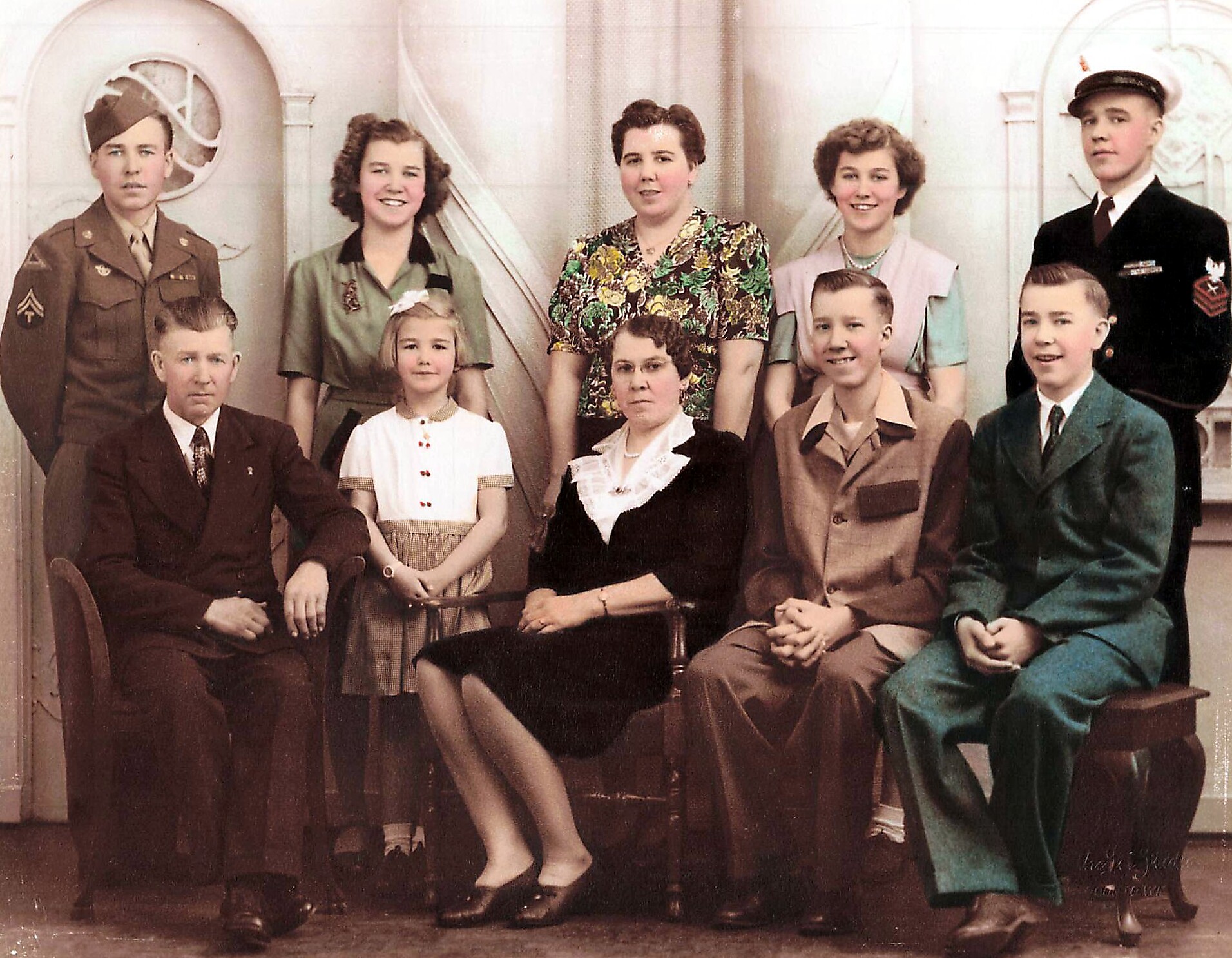 Group portrait of eight people in mid-20th-century attire, some in military uniforms, posing in a studio backdrop.