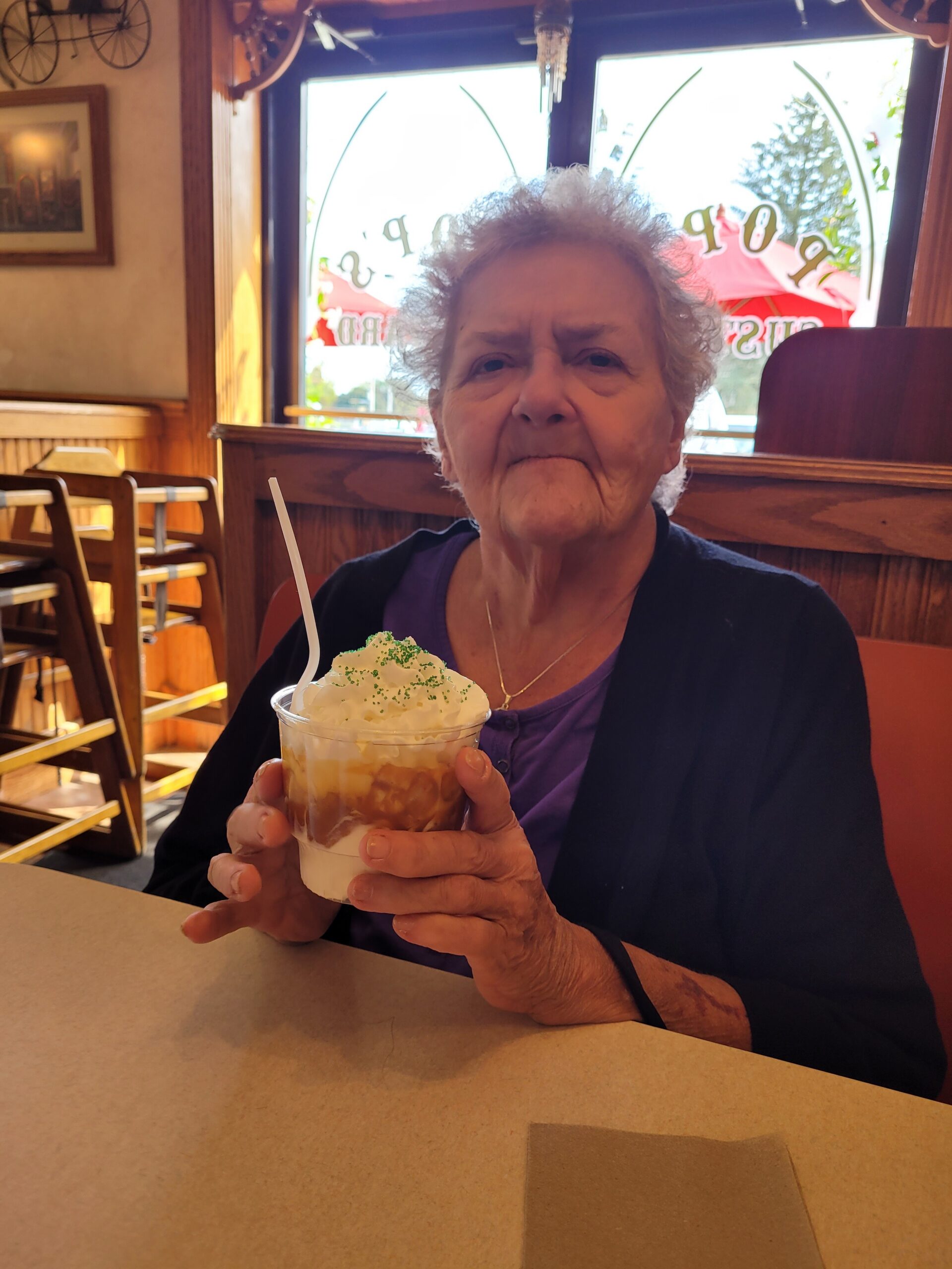Elderly woman in a purple top holds a whipped-cream topped dessert cup with a straw at a cafe.