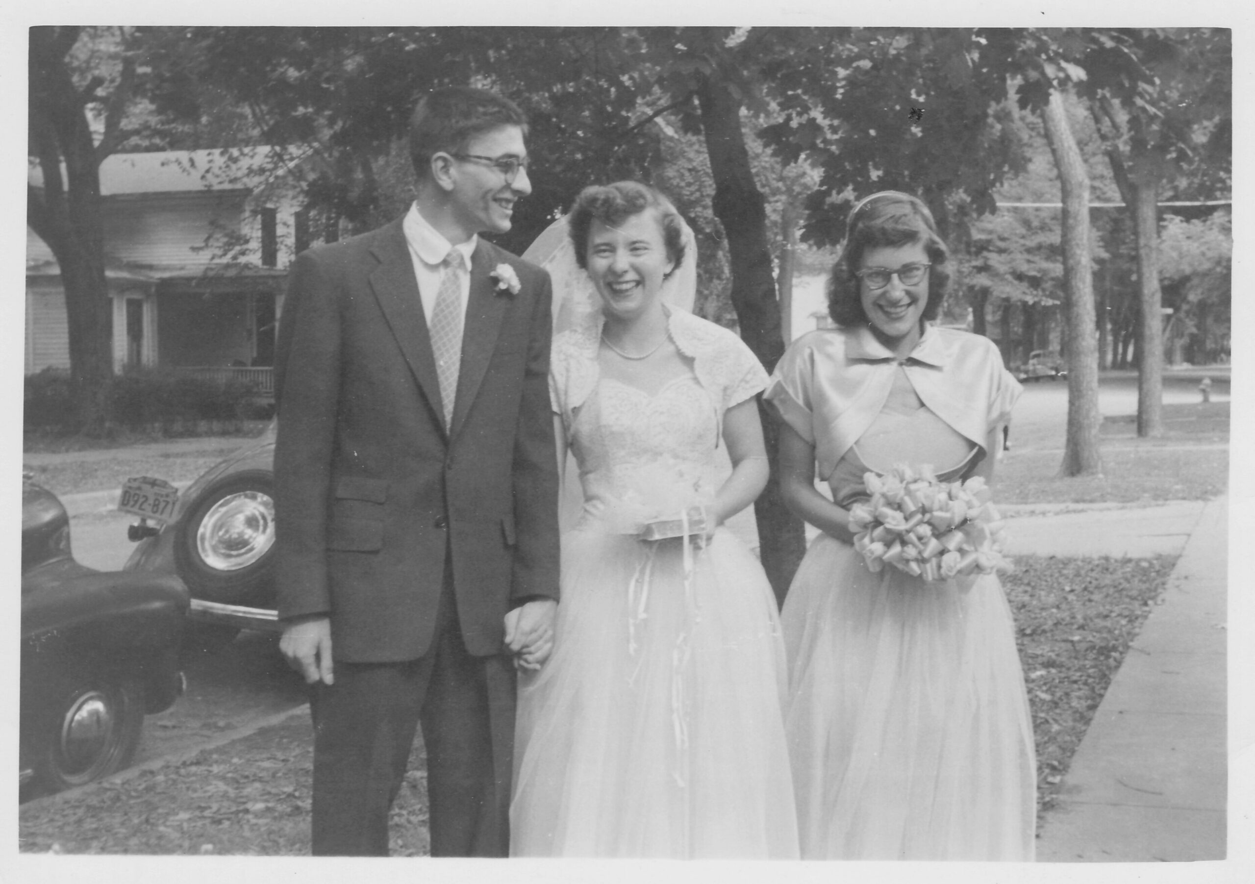 Bride in lace dress and veil walks arm-in-arm with a suited groom while a bridesmaid with a bouquet smiles beside them outdoors on a tree-lined street (black-and-white photo).