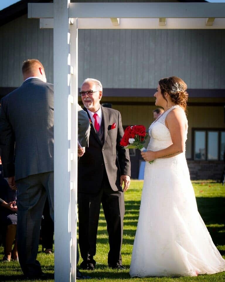 Bride in a white wedding dress holds a red bouquet while two men in suits stand under a white outdoor arch during a ceremony, with guests nearby.