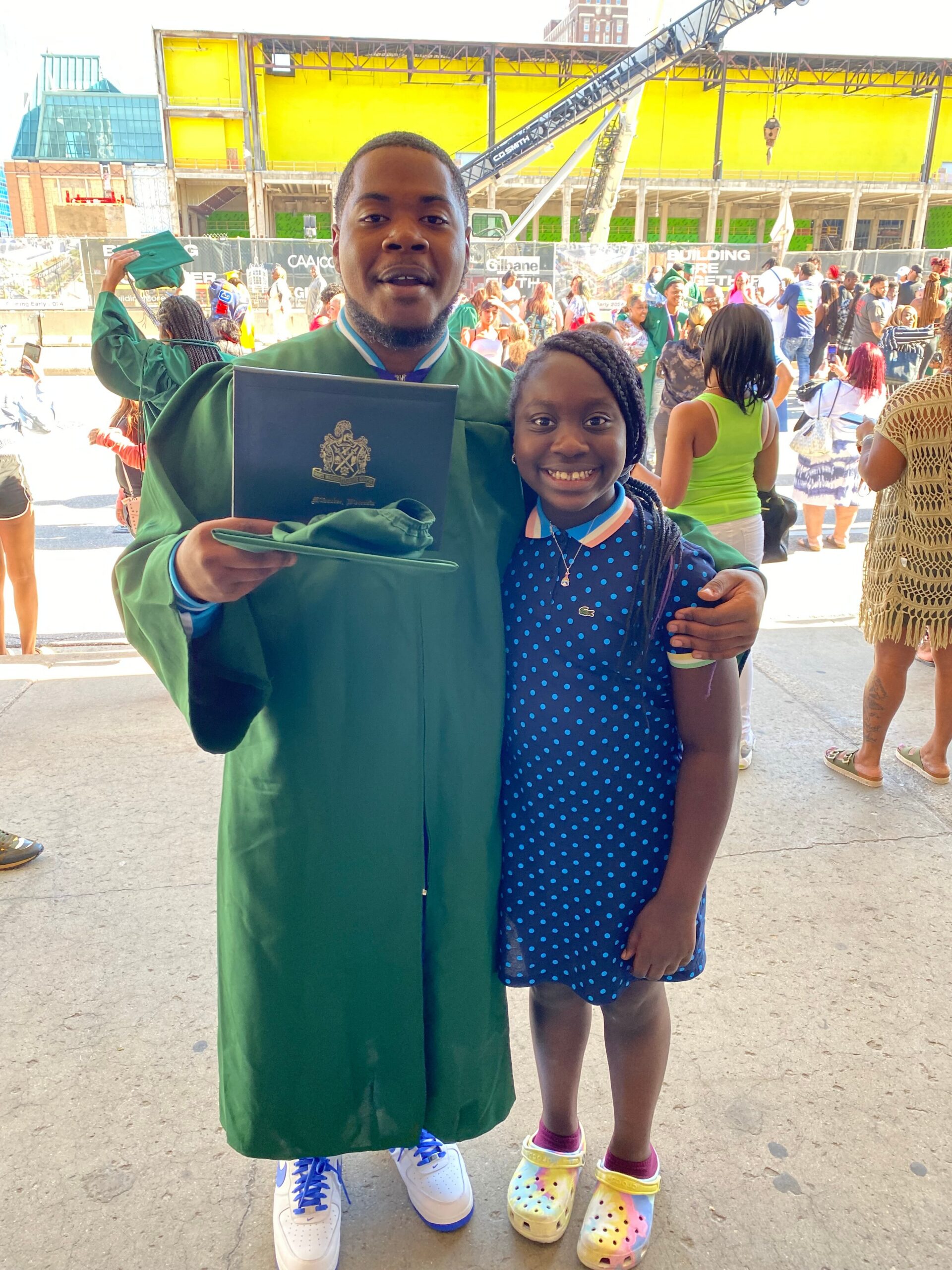 Young man in a green graduation gown holding a diploma and arm around a smiling girl in a blue polka-dot dress at an outdoor graduation celebration.