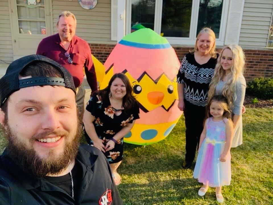 Six people pose for a selfie in a front yard with a giant colorful Easter egg decoration between them.