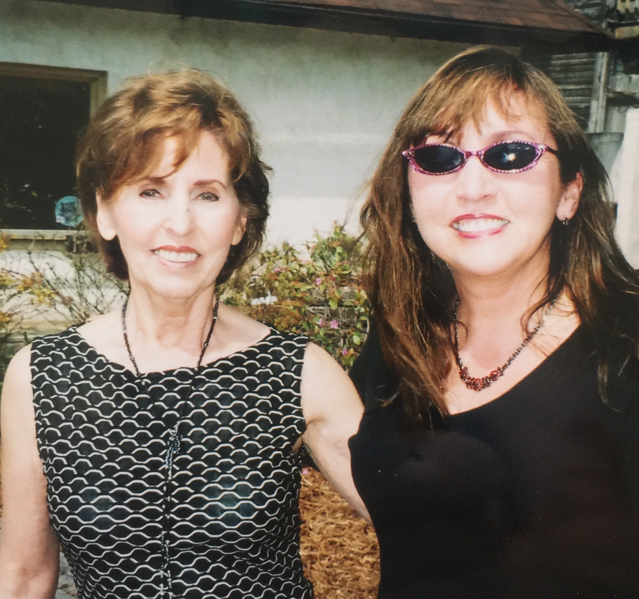 Two smiling women posing outdoors for a photo; the one on the left wears a black-and-white patterned dress, the one on the right wears pink sunglasses and a black top.
