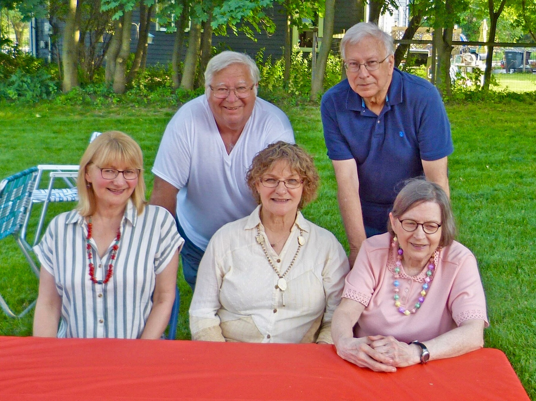 Group of five seniors posing outdoors around a red table in a sunny park setting.