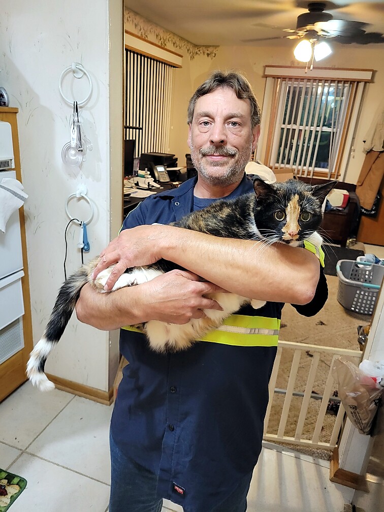Smiling man in navy scrubs holding a calico cat indoors, in a home with kitchen and office area in the background.