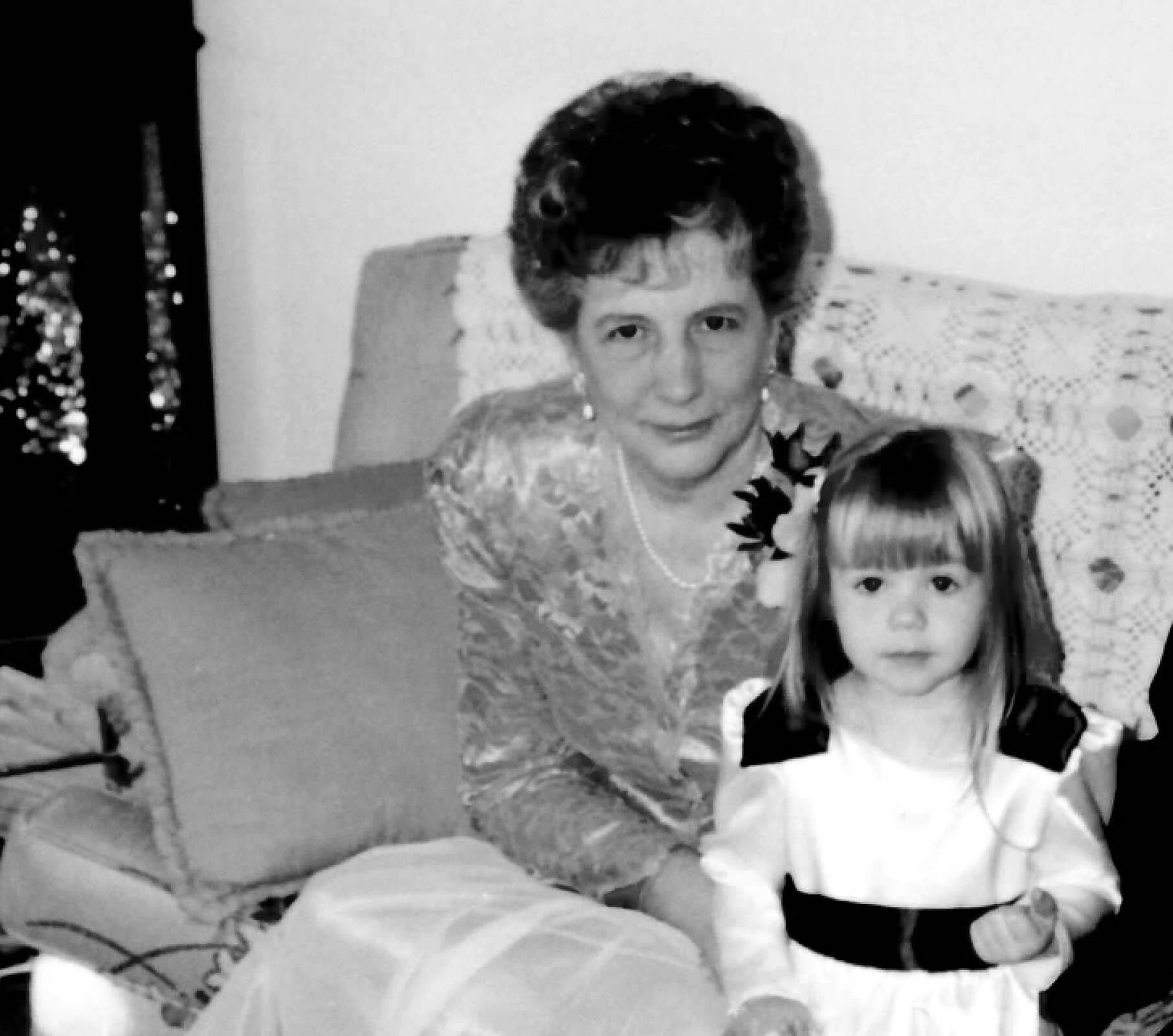 Grandmother with short dark hair in a floral dress sits on a sofa beside a young girl with long hair; both face the camera in a black-and-white photo.