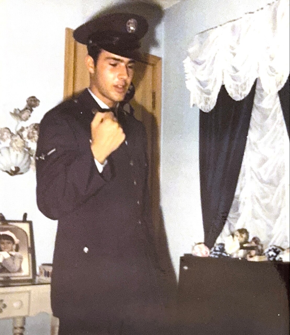 Man in a dark uniform with a peaked cap adjusting his tie indoors, standing near a doorway and a dresser with framed photos.