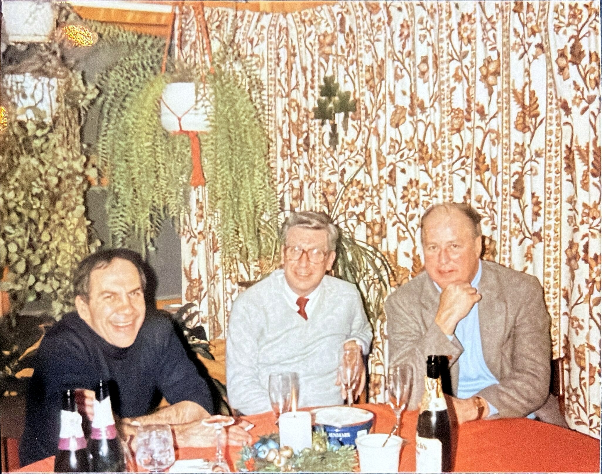 A group of men seated around a table with drinks and snacks, smiling for the photo in a room with floral curtains behind them.