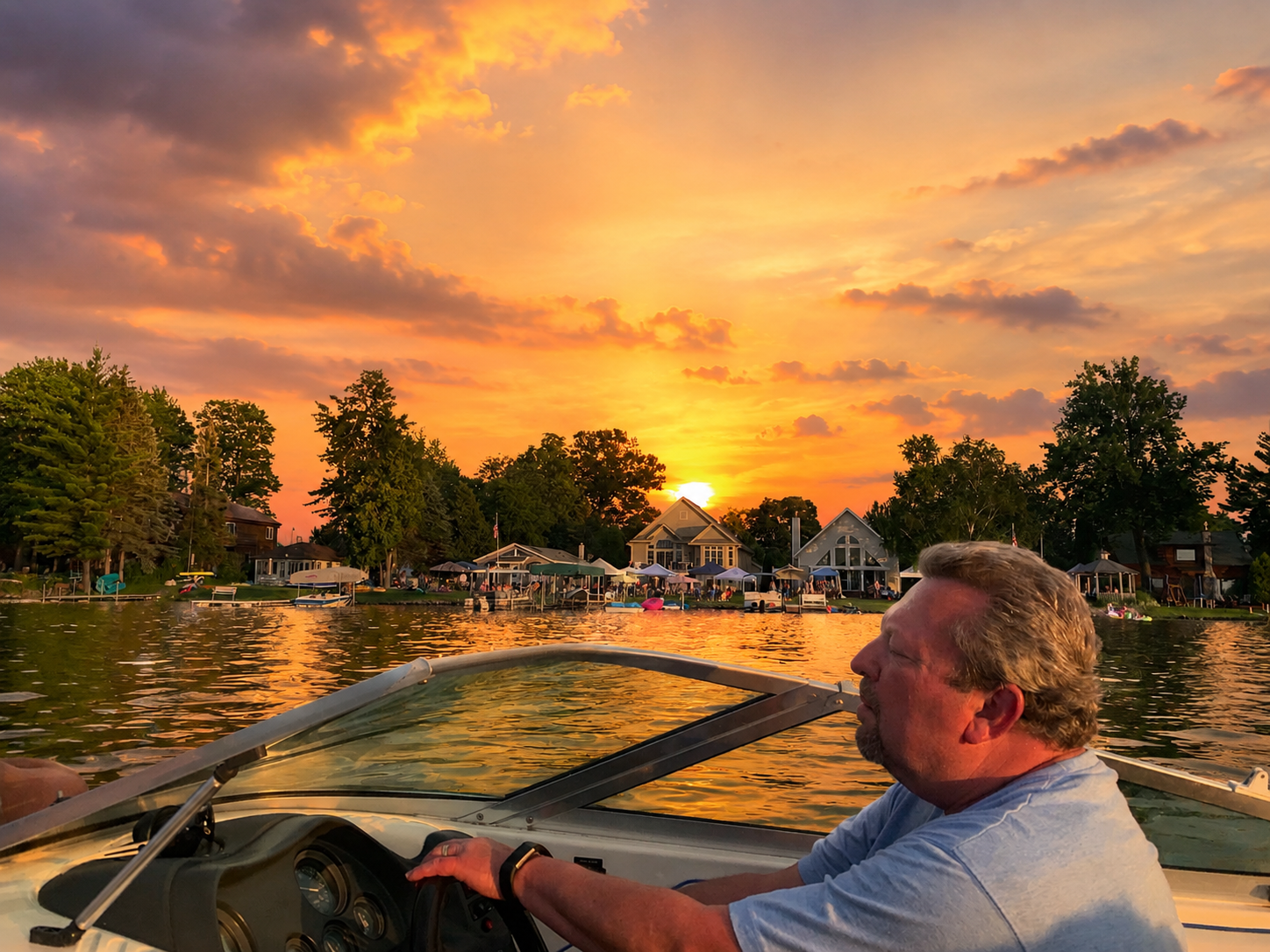 Man steering a boat on a lake at sunset, with a warm orange sky and lakeside homes in the background