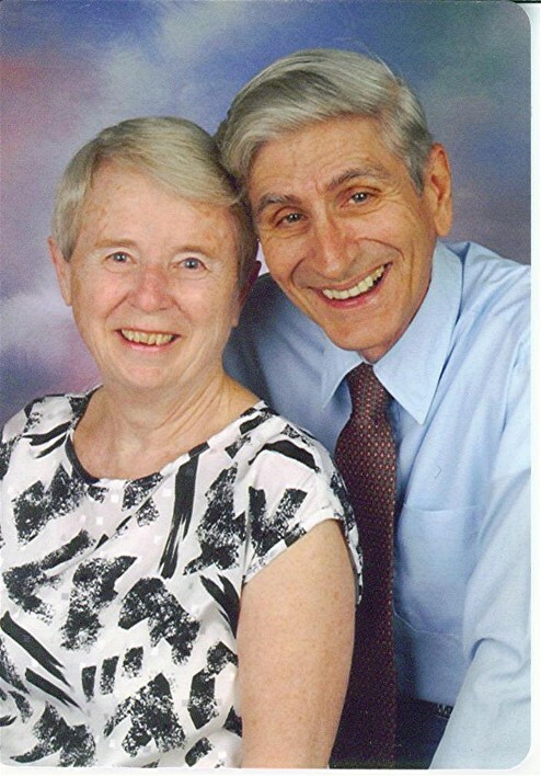 Smiling elderly couple posing closely for a portrait; woman in a black-and-white patterned blouse, man in a blue shirt and maroon tie.