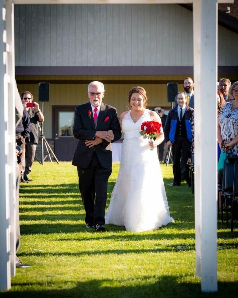 Bride in a white wedding gown holds a red bouquet, walking arm-in-arm with an older man in a suit down a grassy outdoor aisle with guests watching.