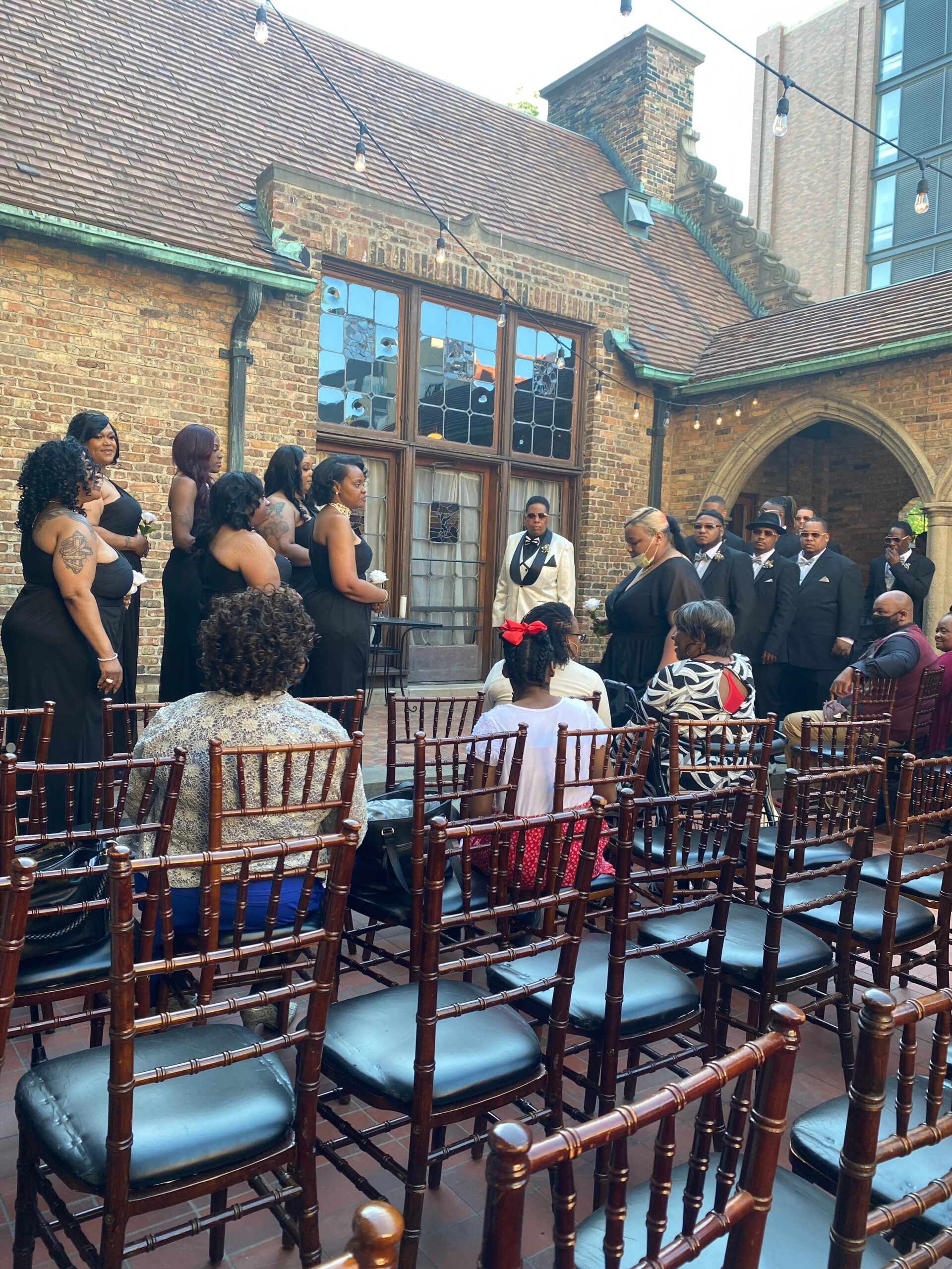 Bridesmaids in black dresses and groomsmen in dark suits stand in a brick courtyard for an outdoor wedding, string lights overhead, wooden chairs set for guests.