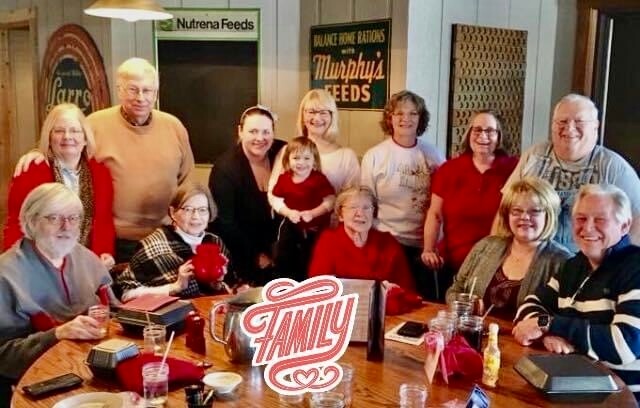 Smiling multigenerational family around a wooden table with drinks and a red 'FAMILY' sign in the foreground.