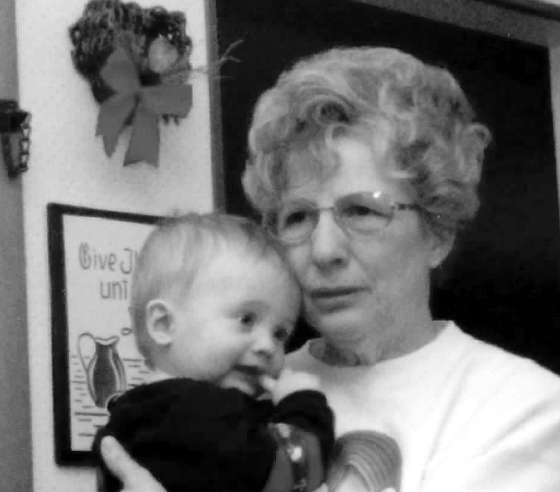 Black-and-white photo of an elderly woman wearing glasses holding a baby while a young child stands in front, near a poster on the wall