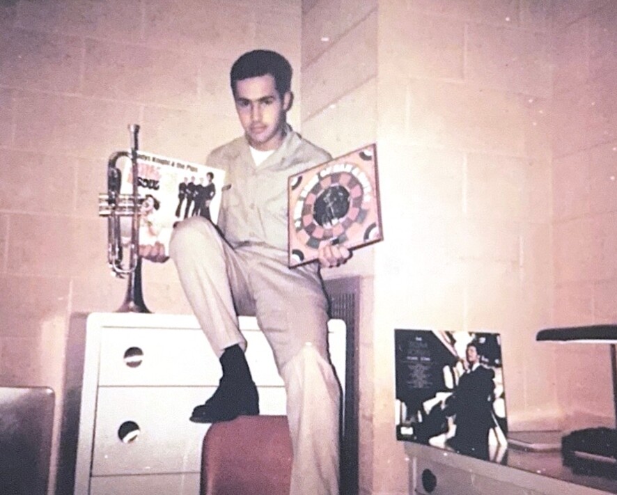 Young man in a beige uniform sits on a dresser, holding a trumpet in one hand and a yellow-framed dartboard in the other, in a retro-styled room.