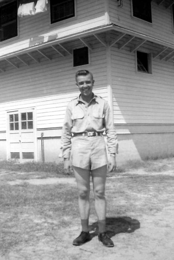 Young man in a light uniform with belt, standing outside a wooden building with a peaked roof and small windows.