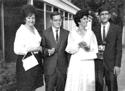 Four adults stand together outside a building, two women in dresses and two men in suits, posing for a group photo.